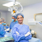 portrait-female-woman-nurse-surgeon-staff-member-dressed-surgical-scrubs-gown-mask-hair-net-hospital-operating-room-theater-making-eye-contact-smiling-pleased-happy-looking-camera-using-trivice
